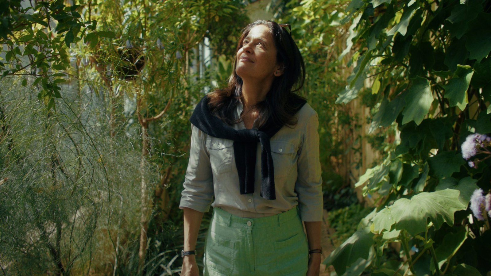 A woman stands in a sunlit greenhouse, looking up at the plants around her
