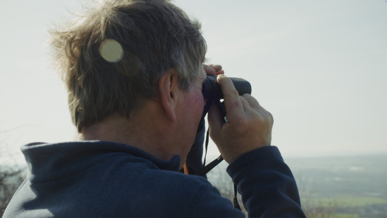 A man looking through binoculars across a hazy landscape, seen from behind