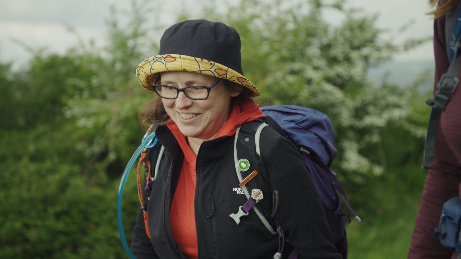 A woman in a sun hat and backpack smiling during a countryside walk