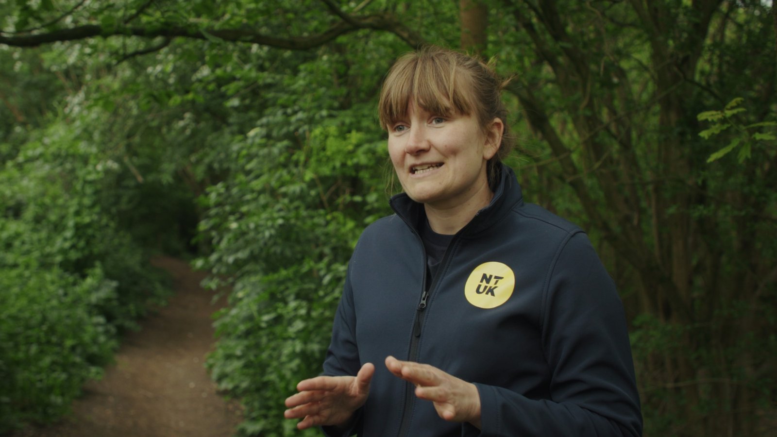 A woman in a National Trails UK fleece gesturing as she speaks on a woodland path