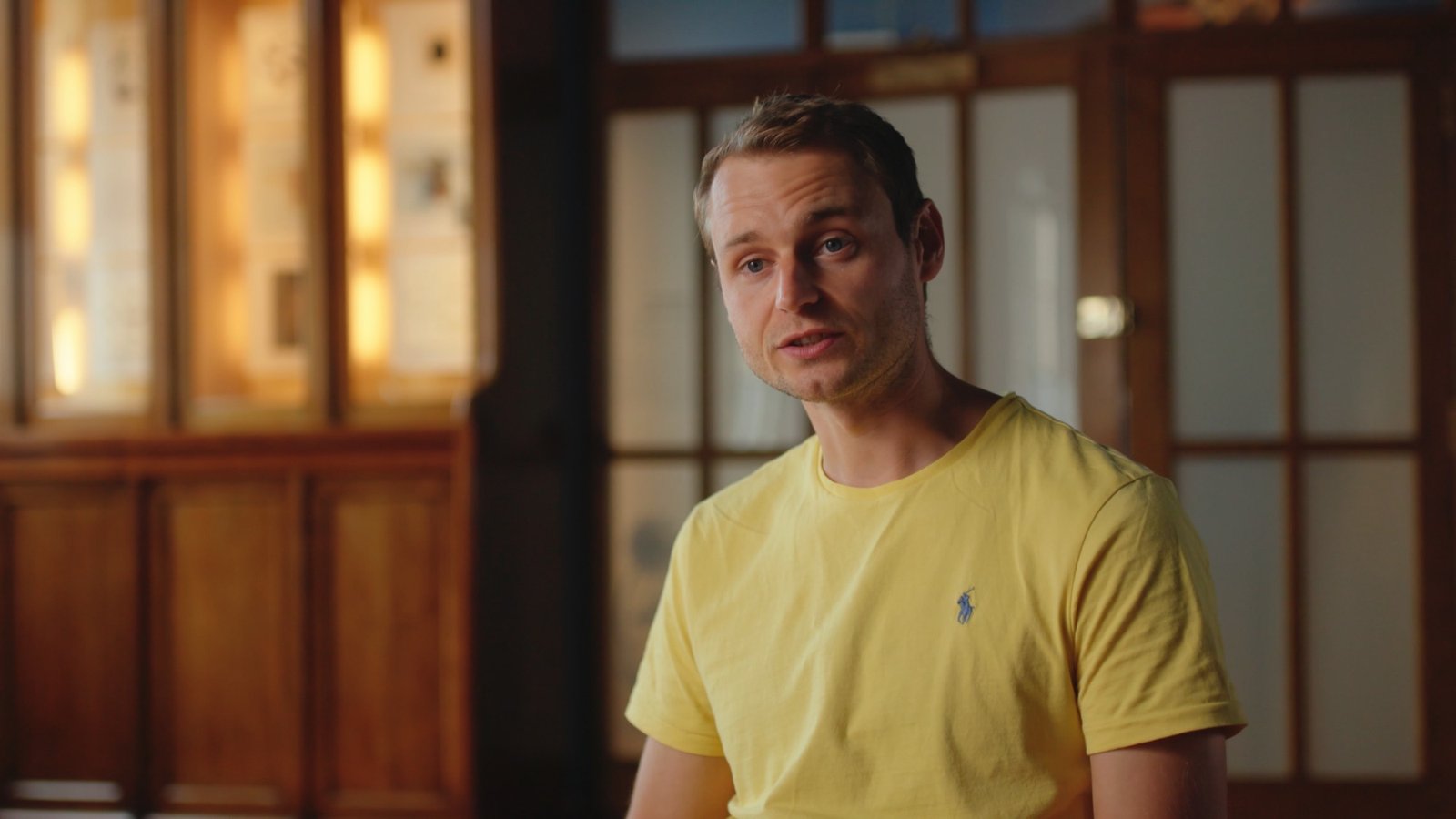 A man in a yellow t-shirt speaking during an interview, wood-panelled room behind him