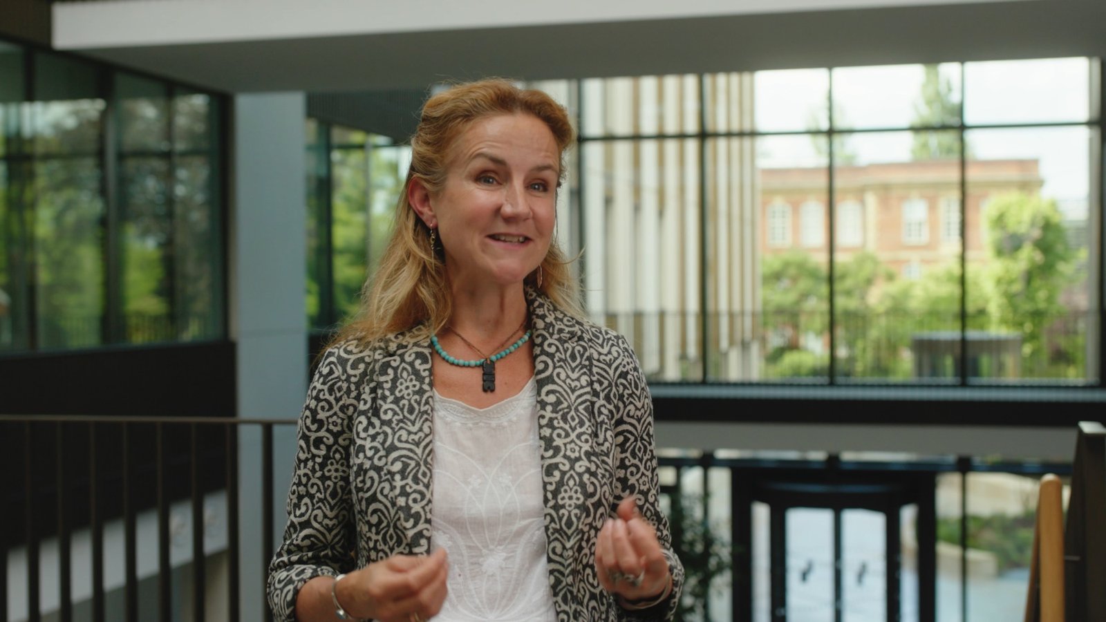A woman in a patterned jacket speaking during an interview in a glass-walled building