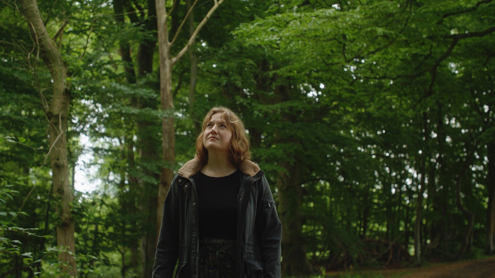 A young woman in a dark coat stands in green woodland, looking upward