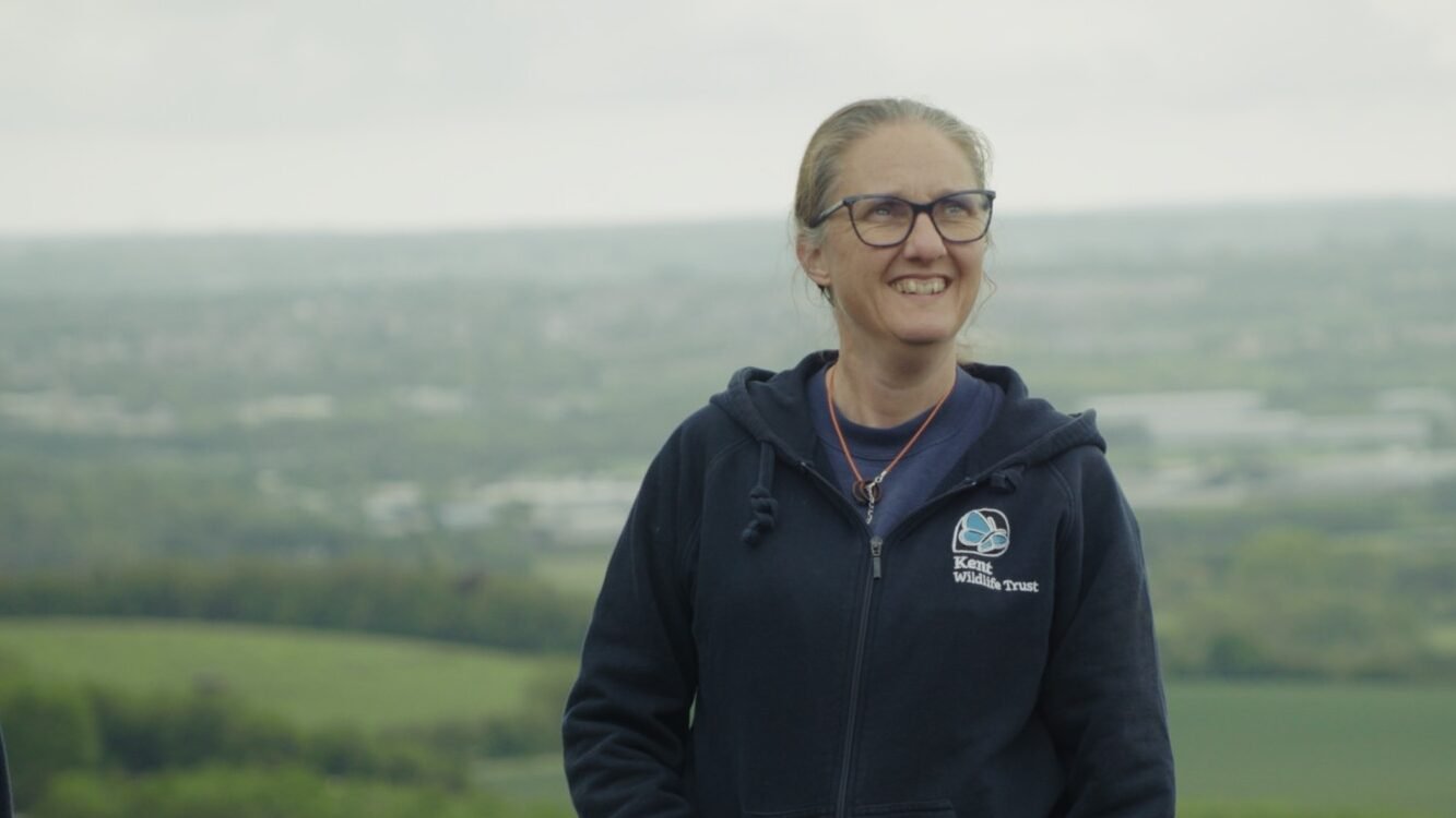 A smiling woman in a Kent Wildlife Trusts hoodie stands on a hilltop overlooking a wide patchwork of fields and woodland