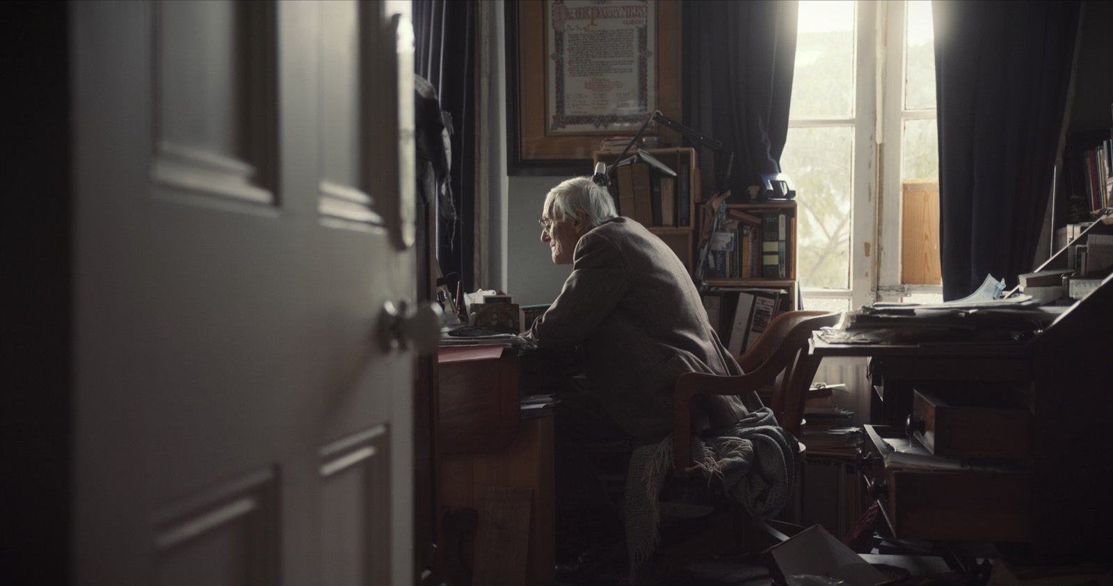 An elderly man with white hair sits writing at a cluttered desk beside a sunlit window