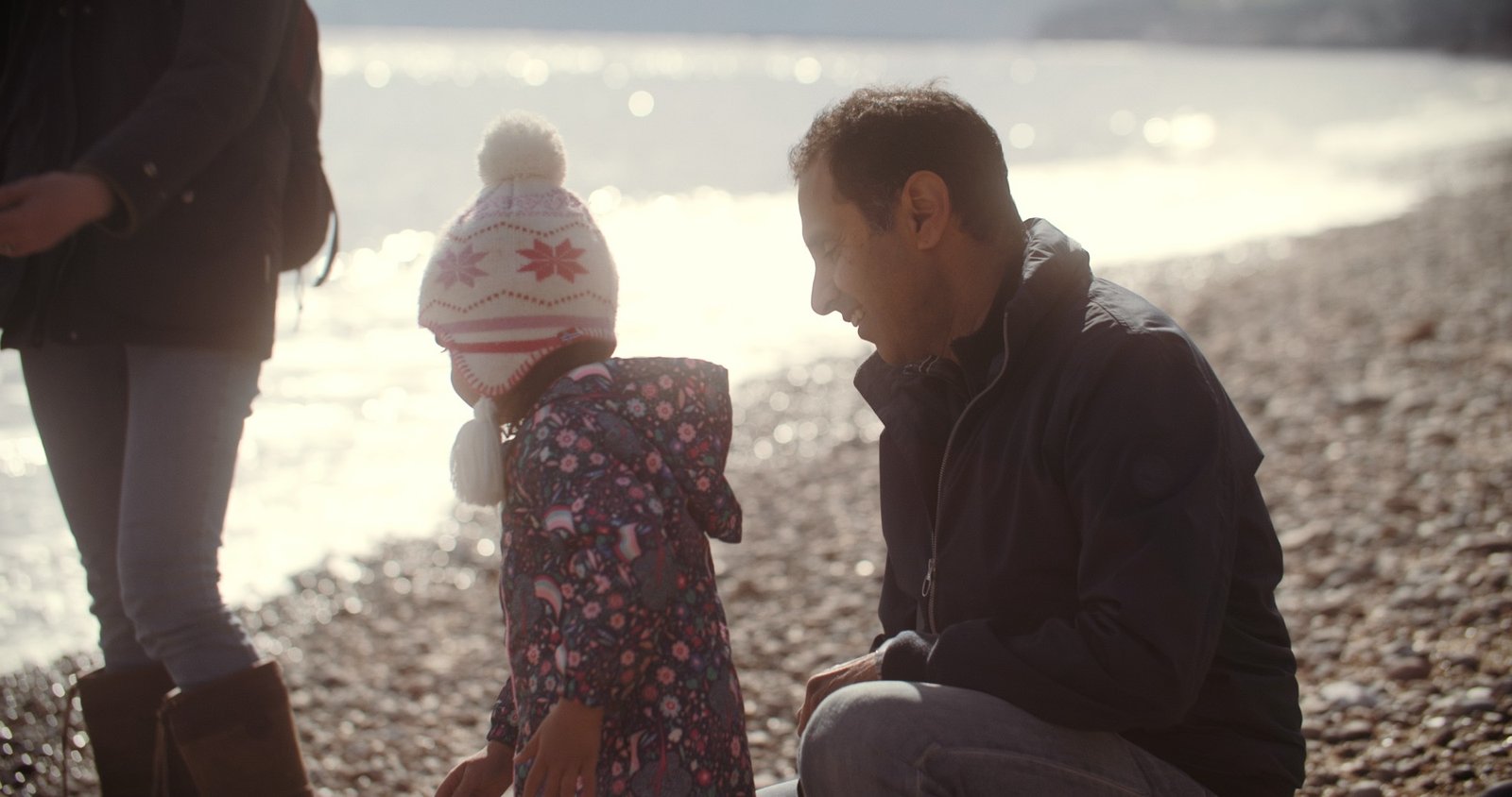 A man crouches to talk with a young child on a pebble beach by the sea
